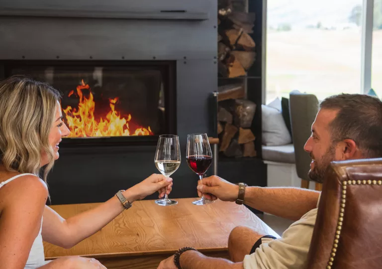 Man and woman smiling in front of a fireplace, holding up glasses of wine