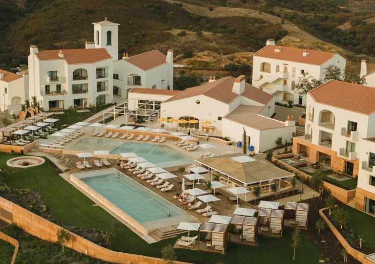 Sky view of white hotel buildings with outdoor pools and tall hills in the background