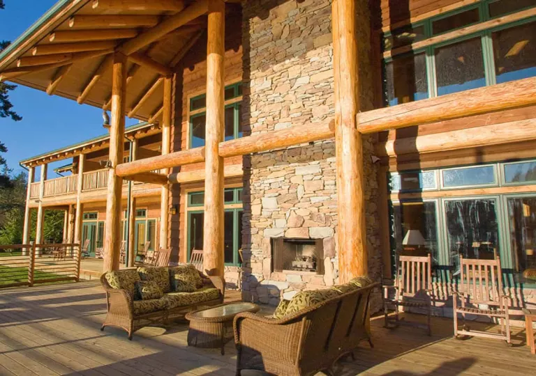 Cabin building exterior with large wooden pillars and a stone fireplace, with cushioned chairs in front