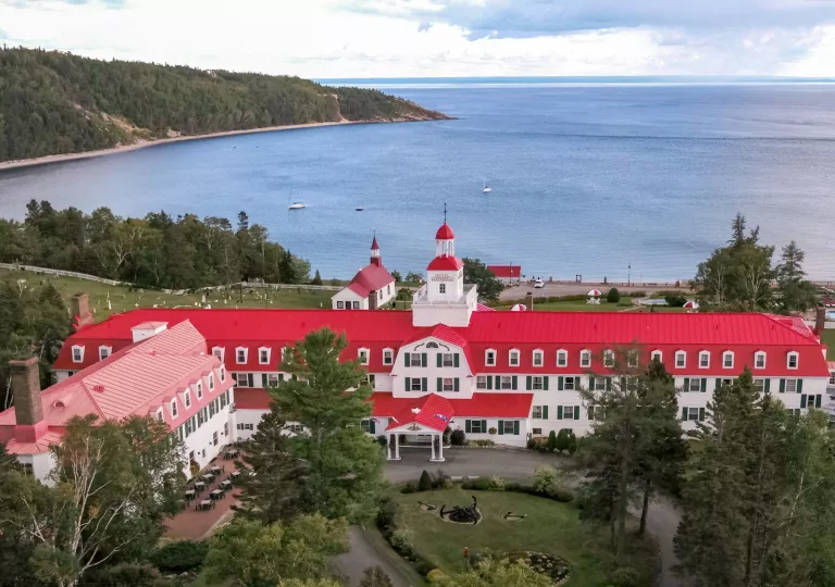 Exterior view of long white building with a red roof, with a large garden in front and the ocean in the distance