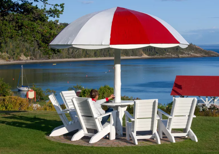 Person sitting on a white lawn chair in front of a red and white table, looking out to the ocean
