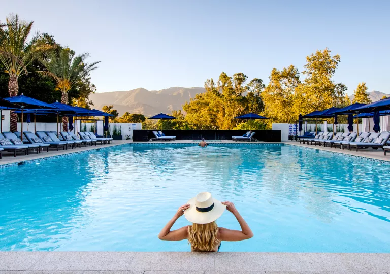 Woman wearing a white hat, standing in an outdoor pool looking towards tall mountains