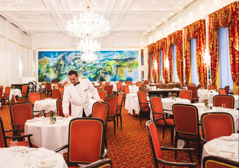 Dining hall with a waiter arranging red chairs around circular white tables
