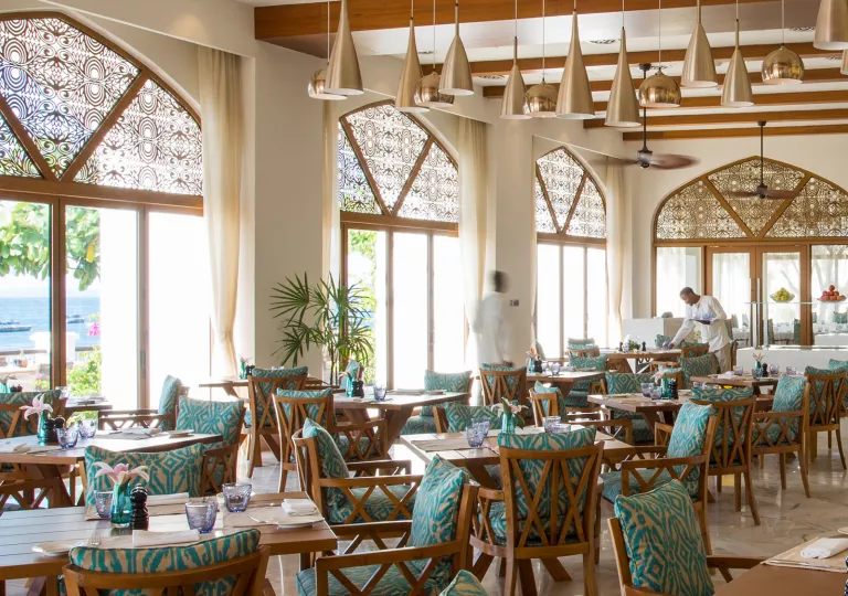 Restaurant dining hall with cushioned chairs surrounding dining tables, with large windows to the left