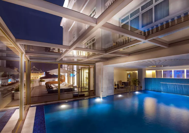 Pool illuminated with lights, in a room with large windows looking out to a hotel balcony