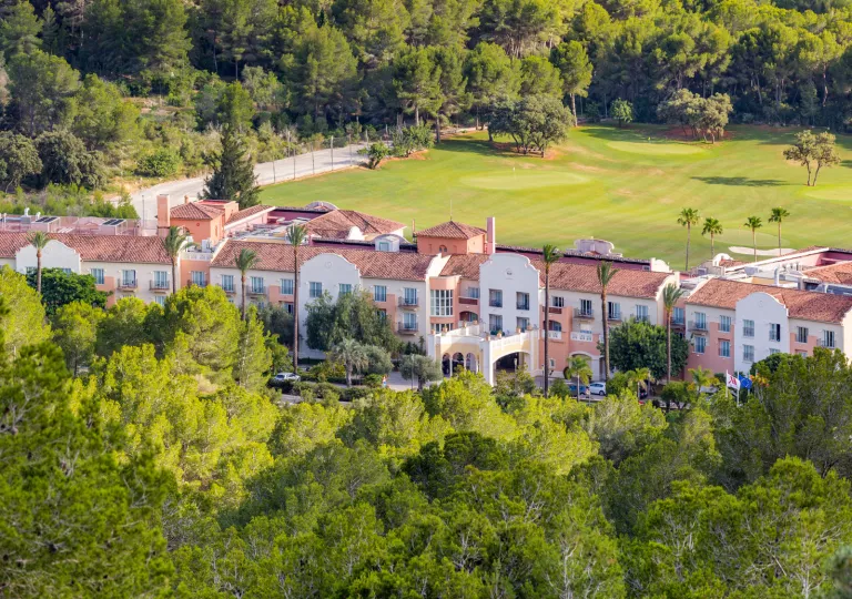 Long, beige and brown hotel building with a large grass field in the back and tall trees in front