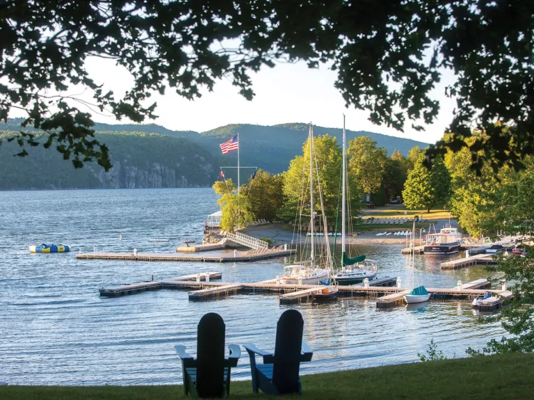Two small, wooden chairs on a field of grass, overlooking a lake docked with boats