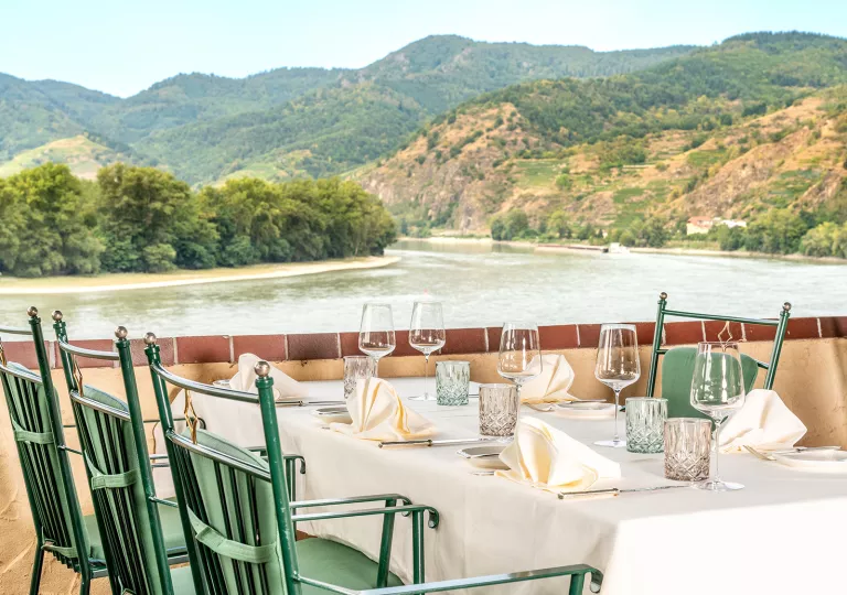 Dining table with green chairs, next to a river surrounded by trees and hills