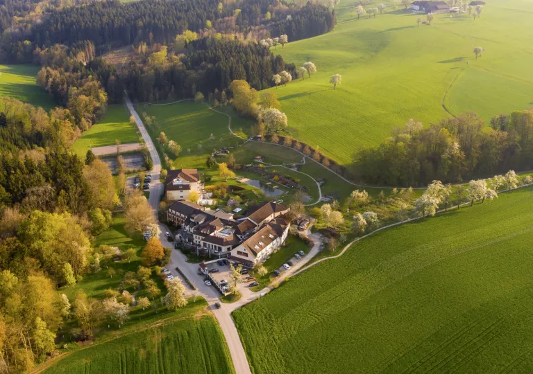 Sky view of large hotel building in the middle of a grassy field, surrounded by a forest