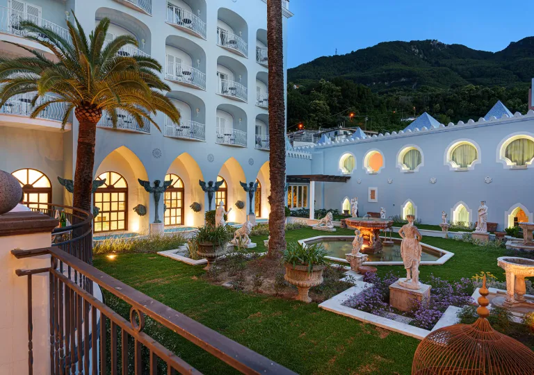 The interior garden of Palazzo Manzi Terme &amp; Spa in Casamicciola Terme on the island of Ischia, Italy features manicured gardens, classical white marble statues, a stone fountain, and large palm trees.
