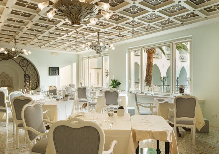 The dining room at the Palazzo Manzi Terme &amp; Spa in Casamicciola Terme on the island of Ischia, Italy features white armchairs, linen-draped dining tables, and a distinctive coffered ceiling accented with decorative chandeliers.