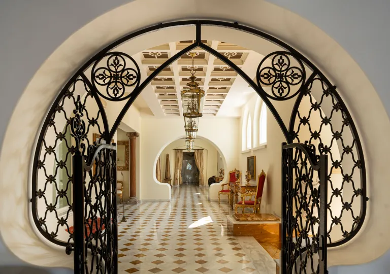 The elegant entryway featuring an arched wrought-iron gate framing a long, bright hallway with a patterned tile floor and classic hanging lanterns at the Palazzo Manzi Terme &amp; Spa in Casamicciola Terme on the island of Ischia, Italy.