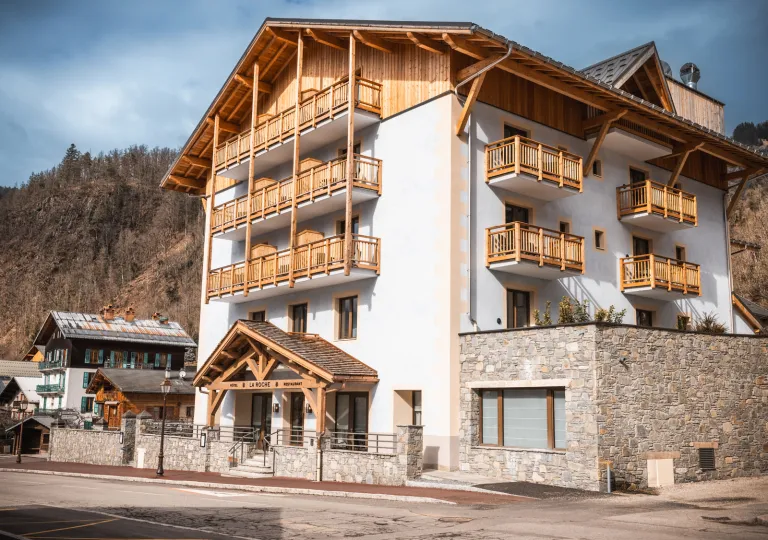 Exterior view of Hotel La Roche, a modern alpine-style hotel with wood balconies, stone accents, and a mountain village setting. The multi-story building is framed by surrounding hills and traditional chalet architecture.