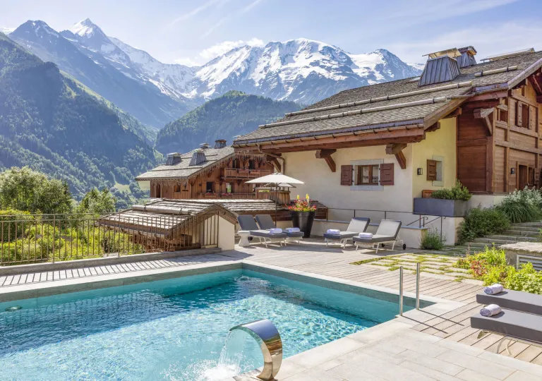 Beige building with an outdoor pool in front, with tall mountains in the distance