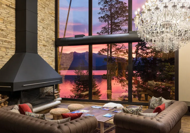 Calfuco Wine Hotel &amp; Spa features a grand, double-height main living lounge framing the shifting sunset colors of Patagonia. The room incorporates woven rattan floor poufs, a solid slab wooden coffee table, plush couches, and an exposed wood pile for the hearth.