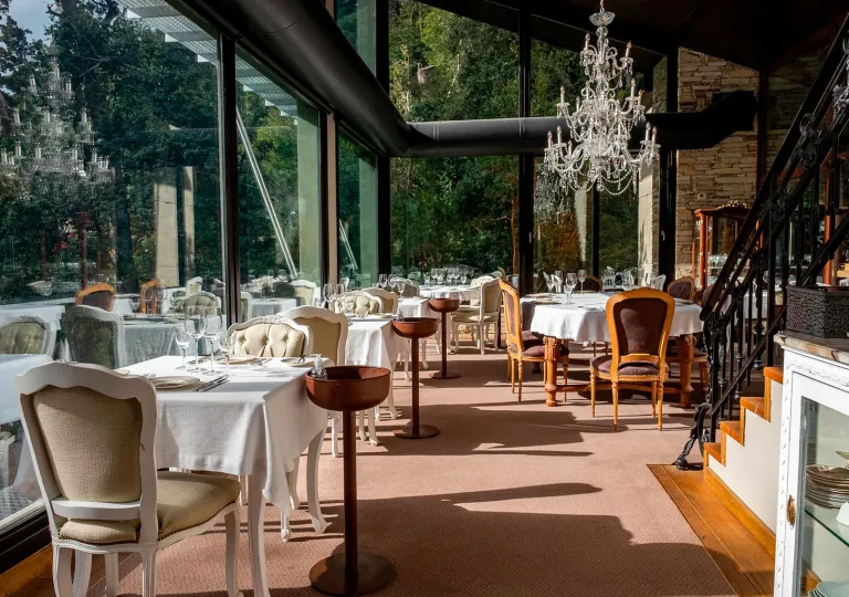 Fine-dining restaurant area inside Calfuco Wine Hotel &amp; Spa featuring white linens, elegant chandeliers, and floor-to-ceiling glass windows that look out directly into the bright green Patagonian forest in Neuquén, Argentina.