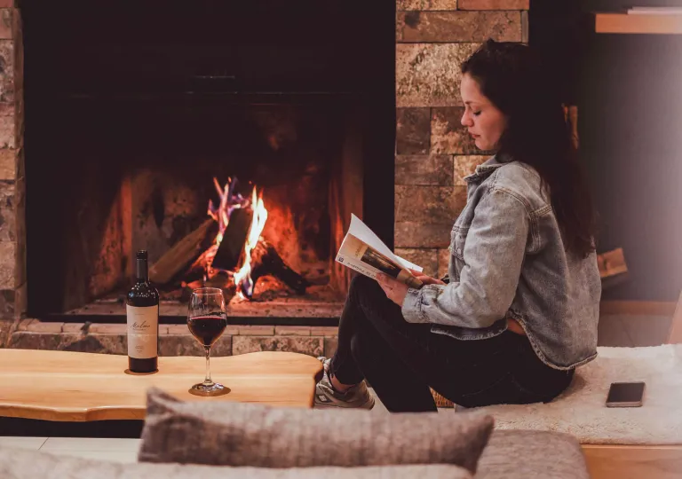 A woman reading near a wood burning fireplace while having a glass of red wine at Hotel Alto Traful.