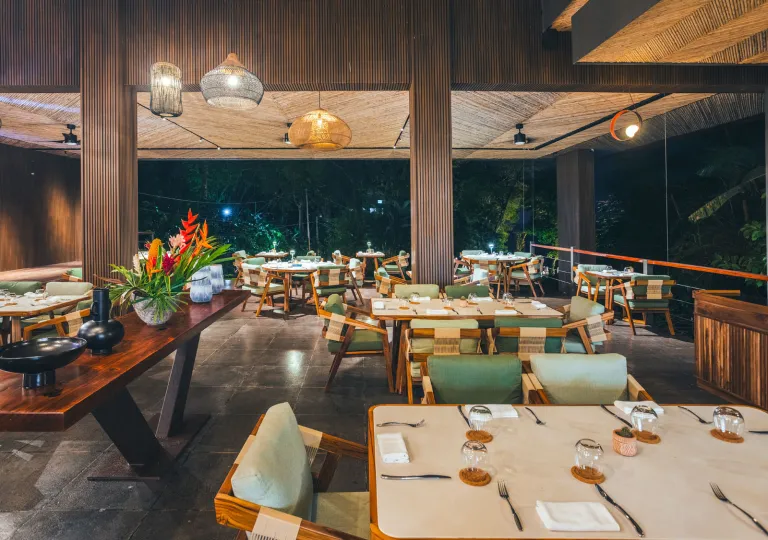 Open-air resort restaurant with wood-paneled architecture, woven pendant lights, and neatly set dining tables. The upscale dining space overlooks lush tropical greenery and is illuminated by warm evening lighting.