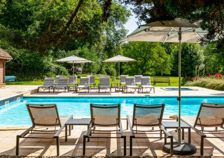 Outdoor pool surrounding by reclining chairs and umbrellas, with large trees in the background