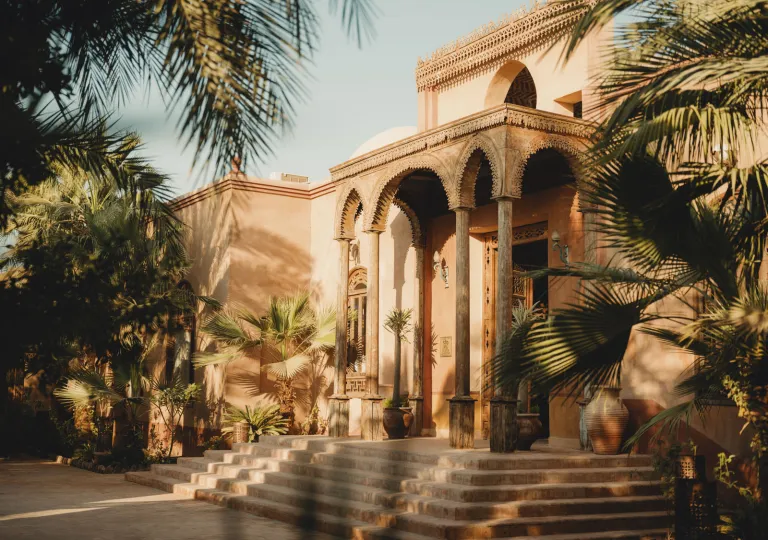 The main exterior entrance of the Al Moudira Hotel in Luxor, Egypt with traditional architecture, featuring hand-carved wooden arches, clay-colored mudbrick structures, and surrounding palm trees.