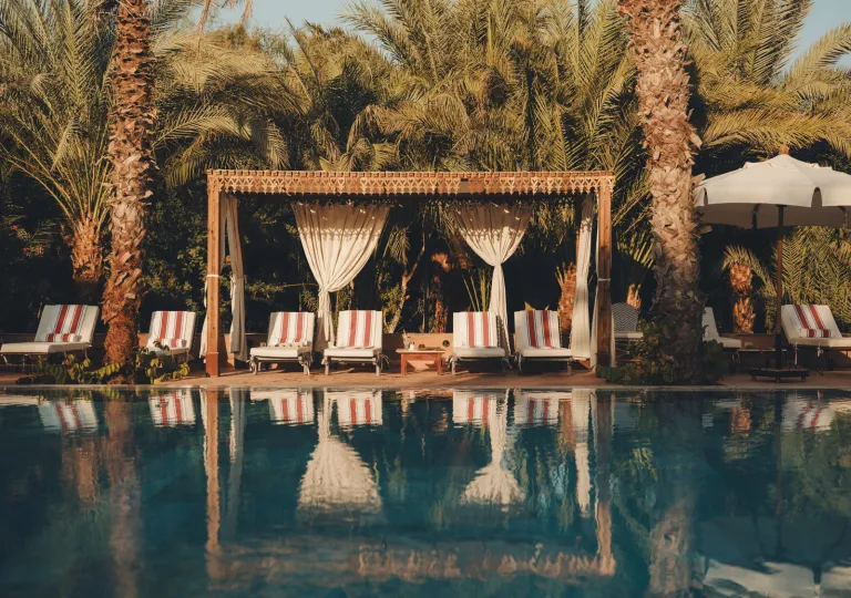 The outdoor swimming pool area at the Al Moudira Hotel in Luxor, Egypt features a large grove of mature date palms, striped lounge chairs, and a hand-carved wooden cabana structure.