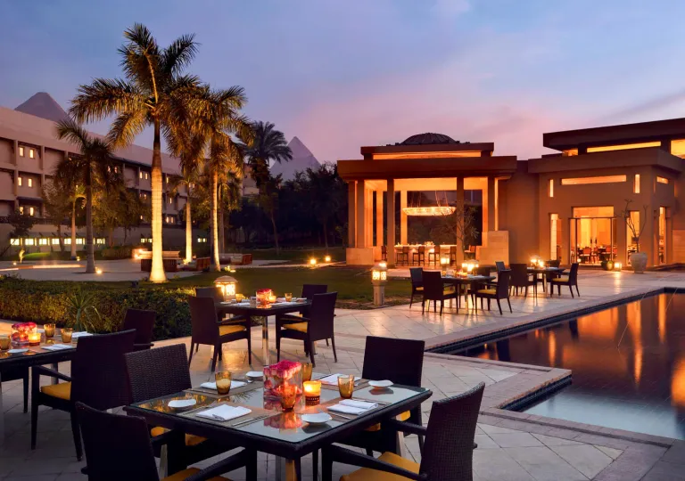 The outdoor dining terrace at the historic Marriott Mena House, Cairo in Giza, Egypt at twilight with the The Great Pyramids of Giza in the background, providing an iconic backdrop for guests dining by the water.