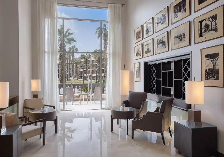 The sun-drenched lounge located inside the Marriott Mena House, Cairo features a wall covered in framed, vintage black-and-white photographs showcasing historical guests, events, and early views of the nearby Pyramids.