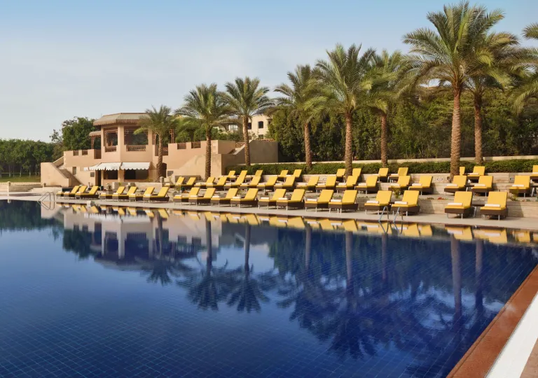 The main swimming pool area at the Marriott Mena House, Cairo in Giza, Egypt features rows of sun loungers with bright yellow cushions that line the multi-tiered stone deck with palm trees overlooking the water.