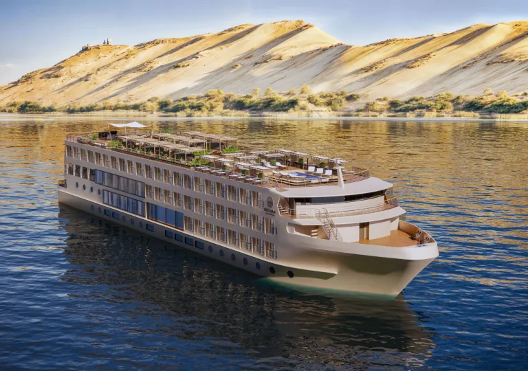 Luxury river cruise ship sailing on the Nile River, featuring multiple passenger decks, outdoor lounge areas, and a rooftop terrace. Golden desert hills rise along the riverbank in the background.