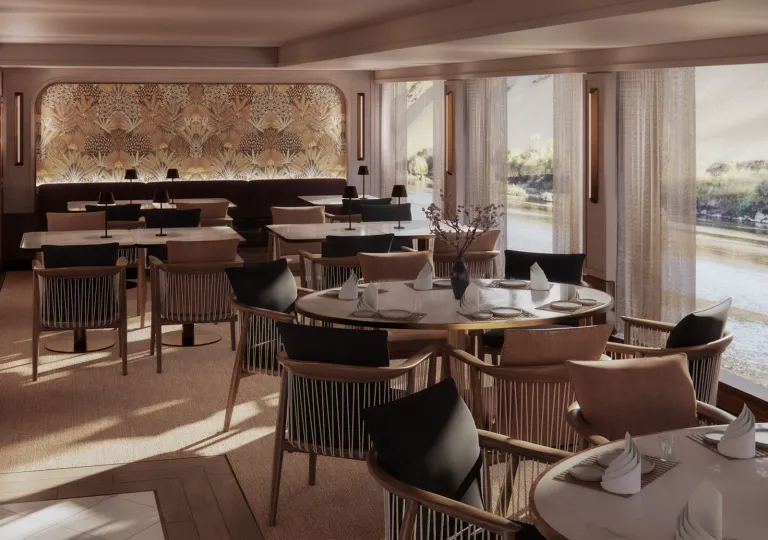 Elegant restaurant aboard a luxury river cruise ship, with round and rectangular tables, upholstered chairs, decorative wall panels, and floor-to-ceiling windows offering views of the Nile River and shoreline.