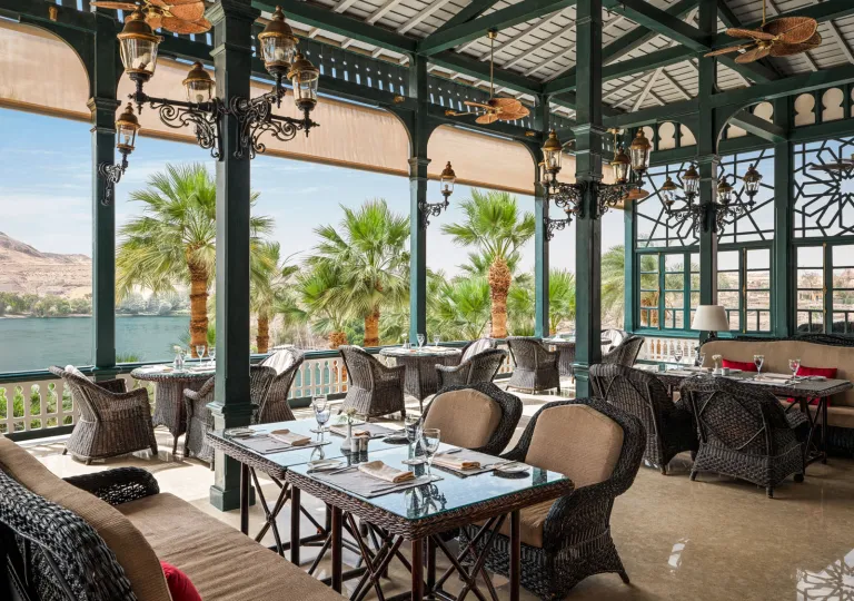 Elegant, open-air terrace restaurant with green wooden pillars, wicker dining tables, and plush seating overlooking the Nile River at the Old Cataract, Aswan in Egypt.