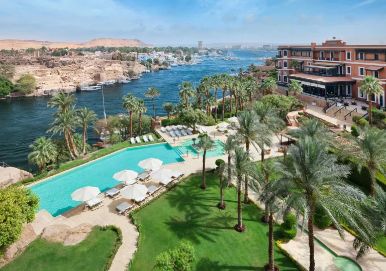 A large infinity-style swimming pool lined with white sun loungers and umbrellas at the Old Cataract Aswan, showcasing its landscaped grounds overlooking the Nile River.