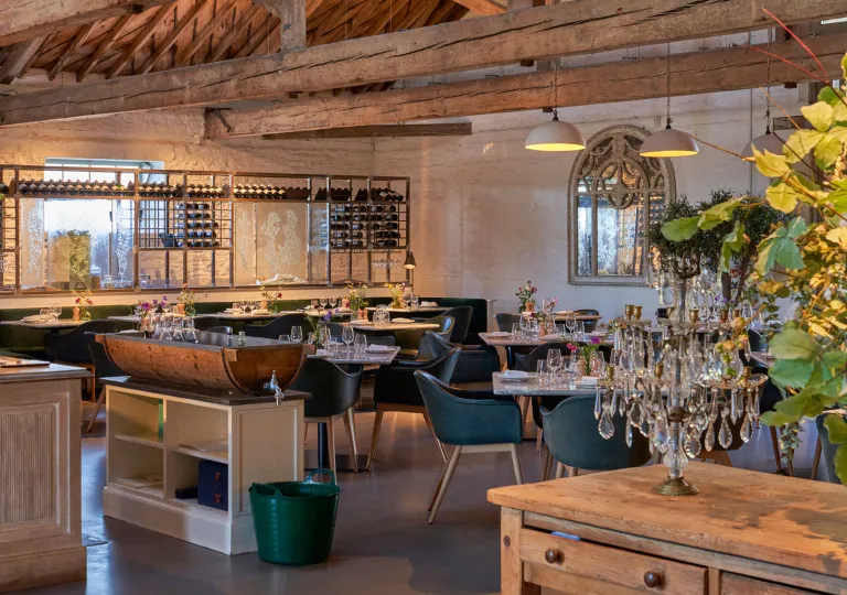 The interior of The Ox Barn restaurant at the Thyme Hotel in the Cotswold village of Southrop, Gloucestershire, United Kingdom that features industrial-style steel wine partitions, arched mirrors, and contemporary dining furnishings.