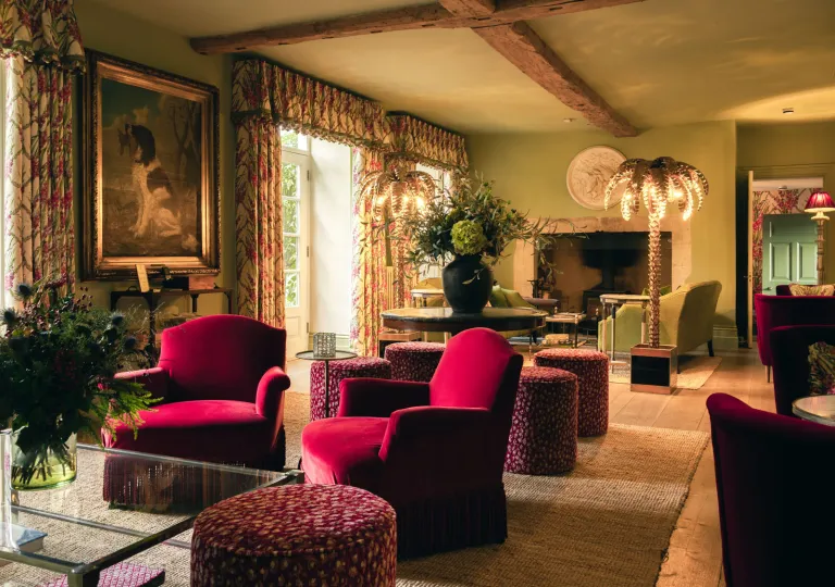 Fifi's Lounge, an elegant drawing room located inside the farmhouse at the luxury Thyme Hotel in Southrop, Cotswolds that features red velvet armchairs and gold palm-tree floor lamps.