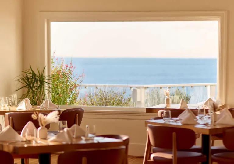 Interior of a restaurant in British Columbia, Canada, with a stunning view of the ocean through a large window. The room is lit with natural sunlight streaming in, casting soft shadows on the wooden floor. Several round wooden tables are set with white tablecloths, folded napkins, and glassware, ready for diners.