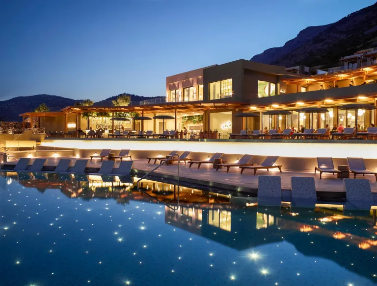 Luxury resort pool area surrounded by tropical landscaping, lounge chairs, and private cabanas. Outdoor dining seating sits beside the pool, with views toward the ocean through the trees in the background.