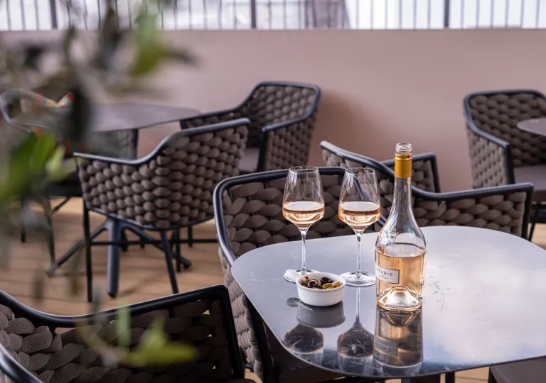 Patio with glass tables and woven chairs, with a bottle of wine and two glasses on a table