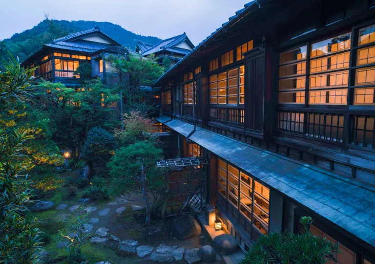 Exterior view of Ochiairo Hotel in Japan at dusk, featuring traditional wooden architecture, illuminated shoji-style windows, and landscaped Japanese gardens set against a forested mountain backdrop.