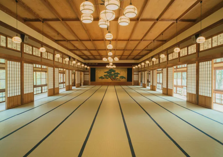 Spacious tatami hall at Ochiairo Hotel in Japan, showcasing traditional Japanese architecture with shoji screens, wood-paneled ceilings, hanging lanterns, and a decorative pine tree mural at the far end of the room.
