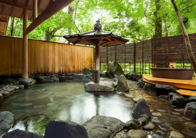 Outdoor onsen at Ochiairo Hotel in Japan, featuring a stone-lined hot spring pool surrounded by natural rock elements, bamboo screens, and lush forest greenery. A traditional wooden shelter and soaking tub create a serene setting immersed in the Japanese countryside.