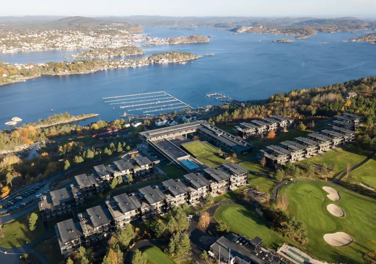 Sky view of buildings in a row, with a golf course in front and a lake in the back