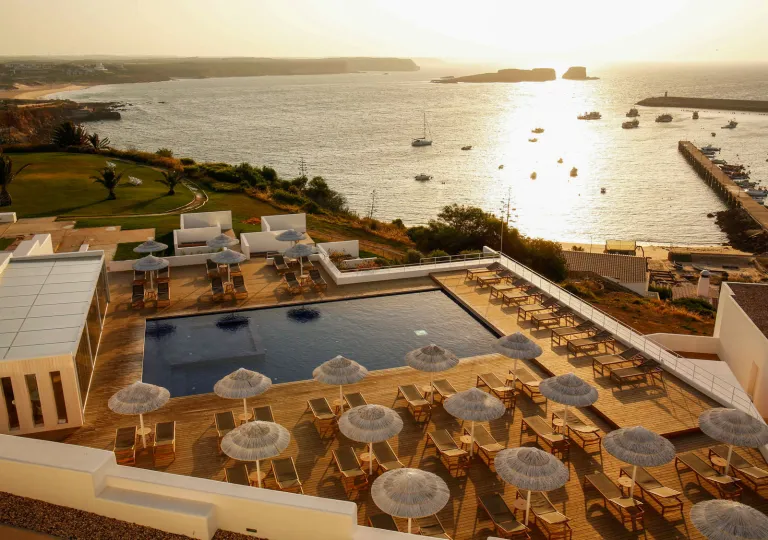 The pool deck area at Memmo Baleeira Hotel in Sagres, Portugal, features a modern, minimalist design and white umbrellas with panoramic views of the Atlantic Ocean.
