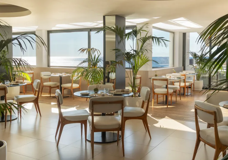 The modern dining area of the Memmo Baleeira Hotel featuring floor-to-ceiling sea views and minimalist design with white cushioned dining chairs and palm tree plants.