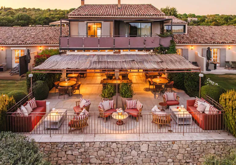A multi-level stone and stucco villa features an outdoor restaurant and lounge setup with several round tables with red striped chairs at Hôtel La Bastide Saint Georges.