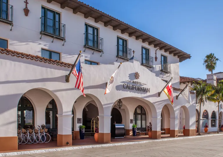 The Hotel Californian in Santa Barbara, featuring Spanish Colonial architecture with a white colored building, arches and flags hanging on the front.