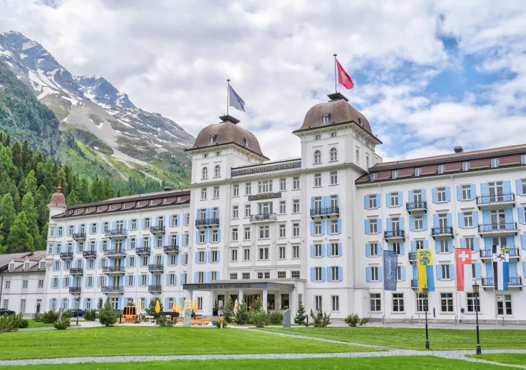 Exterior of the Grand Hotel des Bains Kempinski St. Moritz in Switzerland, featuring a large grand white building with light blue window shutters.