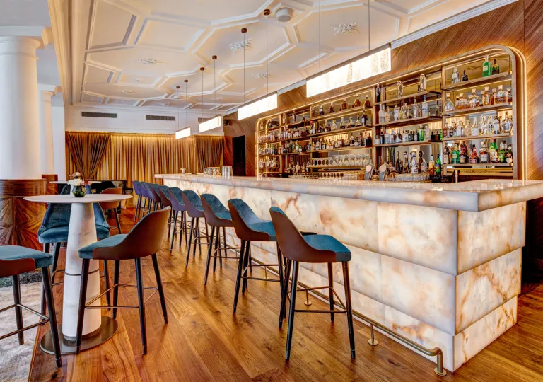 The lobby bar (Quintessence Bar &amp; Lounge) at the Grand Hotel des Bains Kempinski St. Moritz in Switzerland that features a backlit onyx bar, wood-paneled accents and high-backed modern bar stools.