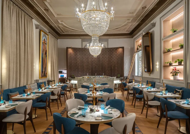 The elegant dining space featuring a tiered crystal chandelier, curved plush velvet seating in deep blue, and warm wooden herringbone floor at the Grand Hotel des Bains Kempinski located in St. Moritz, Switzerland.