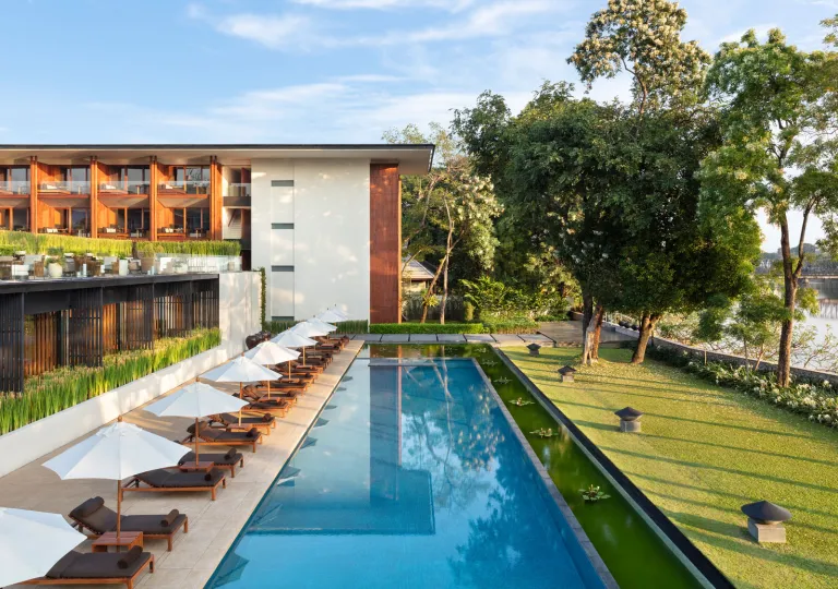 White and brown building with a long, outdoor pool surrounded by chairs and grass fields