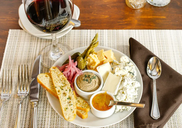 A snack board featuring a variety of curated cheeses, pickled onions, cornichons, a small bowl of pâté or spread, garnished with toasted bread slices and a citrus marmalade.